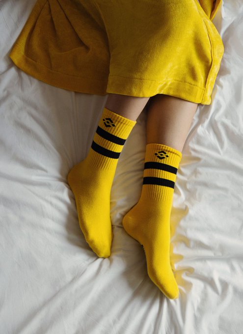 A photo showing a person's legs resting on a white bed, wearing yellow shorts and bright yellow crew socks featuring black stripes and a small black logo near the cuff.
