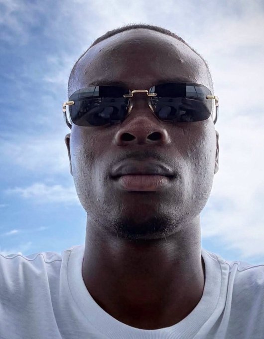 A close-up selfie of a young man wearing dark rectangular sunglasses and a white t-shirt against a blue sky with clouds.