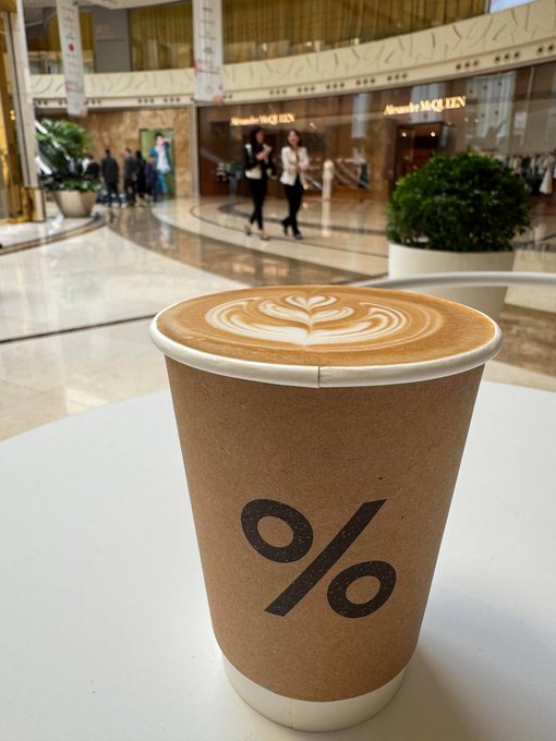 The image features a brown paper coffee cup with a latte art design, placed on top of a white table inside an indoor shopping mall. The cup has a black "%" symbol on its side, indicating that it was purchased using a credit card. A person can be seen walking by in the background, and there are other people visible as well.