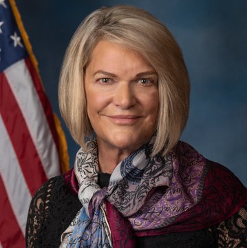 A blonde woman wearing a black lace top and a colorful patterned scarf stands in front of an American flag, smiling at the camera. This appears to be Senator Cynthia Lummis, who is featured in the post advocating for the timely passage of the crypto CLARITY Act.