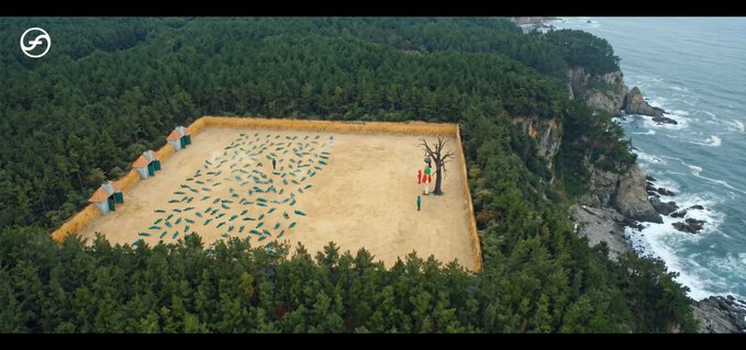An aerial view of an outdoor art installation or performance setup on a clifftop overlooking the ocean, showing a rectangular sandy area with scattered geometric objects, a bare tree, and figures holding colored flags.