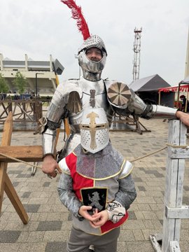 A person in medieval knight armor with a red plume helmet posing with another person wearing a matching tunic, at what appears to be a Renaissance fair.