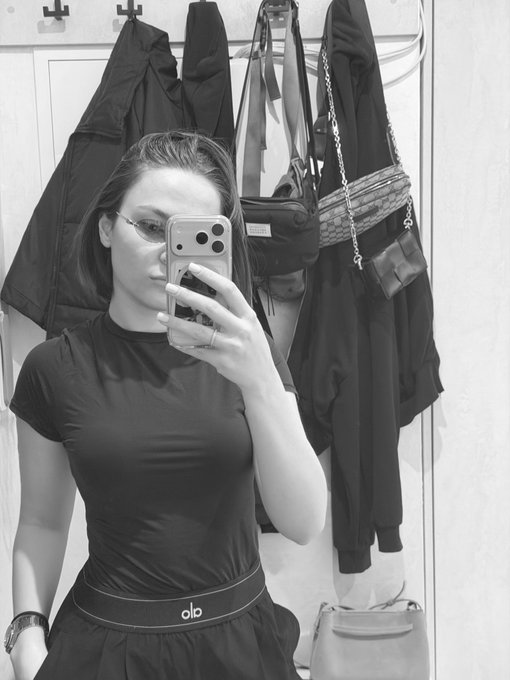 A black and white mirror selfie of a woman wearing glasses and a fitted black top with 'alo' visible on the waistband, taking a photo with her smartphone.