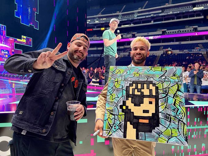 Two men pose at an arena event; one holds up a colorful pixel-art style painting of a Minecraft character.