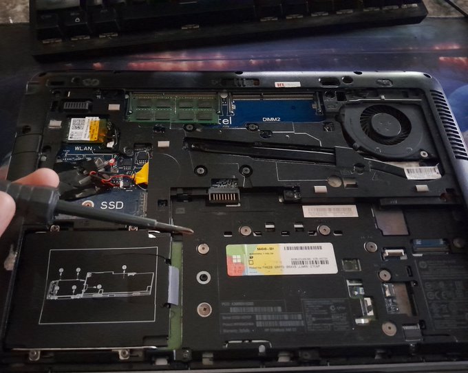 The exposed internal components of a disassembled laptop, showing the SSD, RAM, cooling fan, and a Windows 8 sticker.