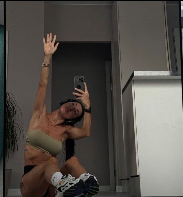 A mirror selfie of a woman wearing headphones and athletic wear, stretching her arm upward in a modern interior.