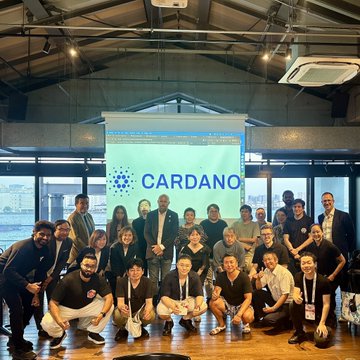 A group photo of people posing in front of a projected 'CARDANO' logo at what appears to be a corporate or community event.