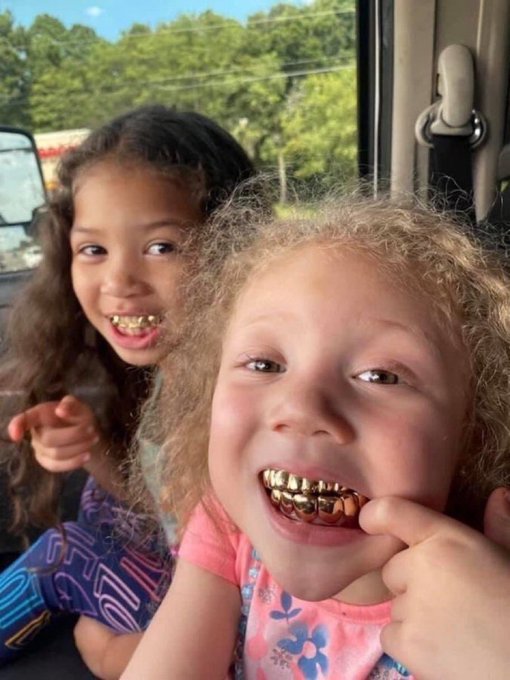 A close-up photo of two young smiling girls inside a car, both wearing shiny gold grillz on their teeth, with green trees visible through the car window in the background.