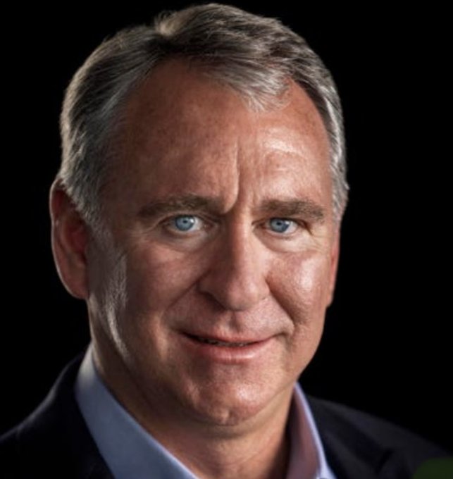 A professional headshot of a middle-aged man with gray hair and blue eyes, smiling slightly against a black background.