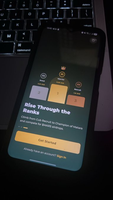 The image shows a cell phone with an app called "Rise Through The Ranks" displayed on its screen, which appears to be related to a loyalty program or a mobile game. The phone is placed next to a laptop computer that has a keyboard visible in front of it.