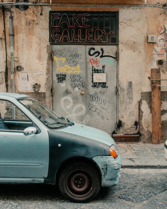 A street scene featuring an old light blue compact car parked in front of a weathered building with a metal door covered in graffiti, topped by a red neon 'FAKE GALLERY' sign.