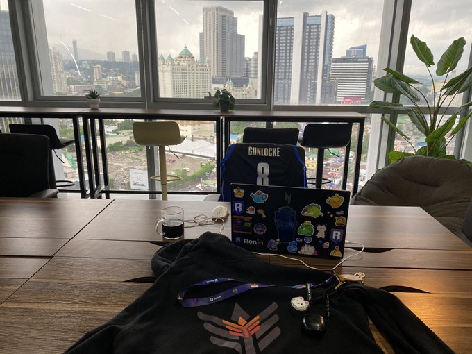 A workspace setup with a wooden desk, a laptop covered in stickers (including 'Ronin'), a jersey with 'GINLOCKE' and the number 8, and a view of a cityscape through large windows.