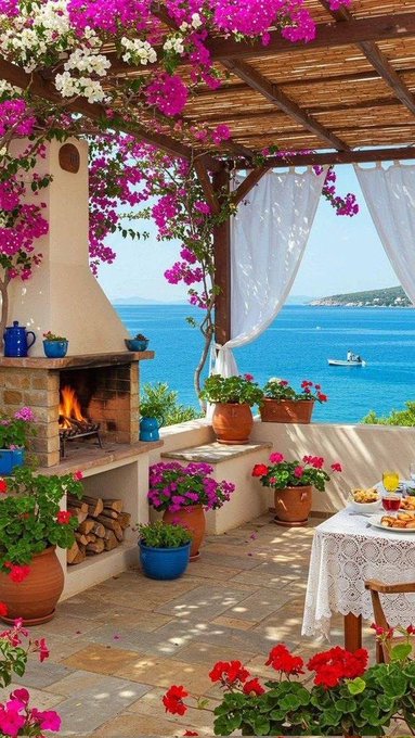 A picturesque Mediterranean-style outdoor patio features a stone fireplace, lush pink and purple bougainvillea flowers, white curtains, and potted plants overlooking a blue sea with a small boat.