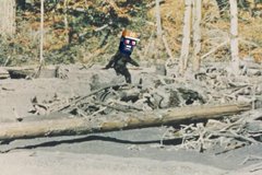 A pixelated image of a small black figure running across a field of debris and logs, with trees in the background.