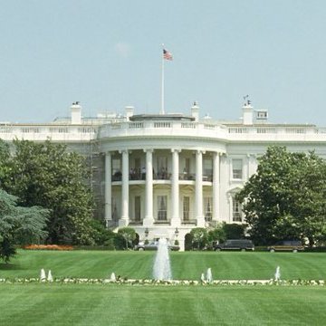 The White House is shown with its iconic South Portico and an American flag flying overhead, with a fountain and lush green lawn visible in the foreground. The image directly relates to the post, which discusses a White House analysis on the impact of banning stablecoin rewards on bank lending.