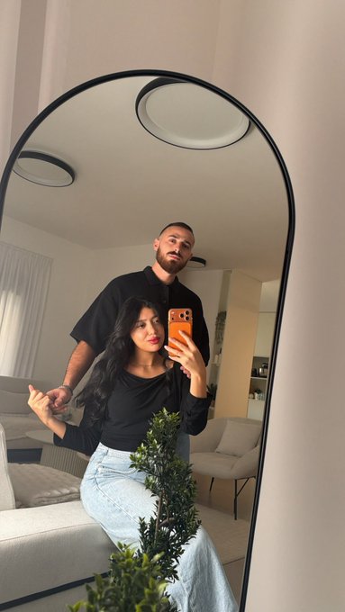 A couple poses for a mirror selfie in a bright living room, with the woman holding an orange-cased phone and a leafy plant in the foreground. No names, numbers, or tickers are visible; the post’s “Best caption gets 1 SOL” text is not shown in the image.