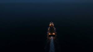 An aerial nighttime view shows a ship traveling through dark waters, leaving a wake of foam illuminated in its path.