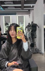 A mirror selfie of a young woman with dark hair and glasses holding a yellow phone in a gray jacket, taken in what appears to be a clothing store.