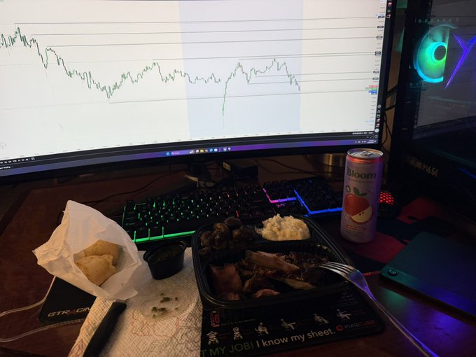 A trader's desk with a monitor displaying a downward-trending stock chart, alongside a meal of meat, mashed potatoes, and bread, plus a can of 'Bloom' sparkling apple cider.