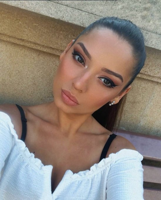 A close-up selfie of a woman with dark hair pulled back, wearing dramatic eye makeup with winged eyeliner, a nose ring, and a white off-shoulder top with thin black straps, posed against a beige textured wall.