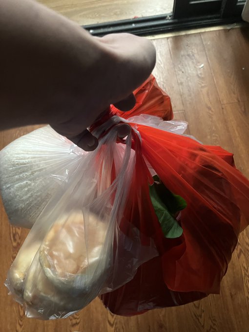 A hand holds up several plastic grocery bags, including a red one, containing food items like bread and what appears to be rice on a wooden floor.