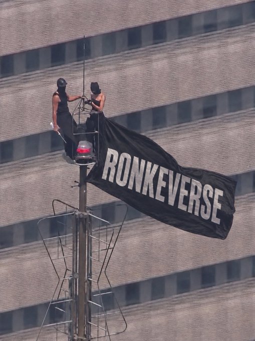 Two people at the top of a tall pole unfurling a large black banner that reads 'RONKEVERSE' against a brick building backdrop.