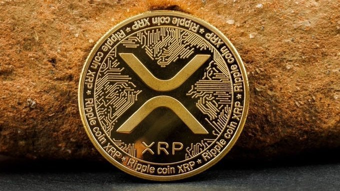 A gold XRP (Ripple) commemorative coin rests on a stone surface, displaying the XRP logo and the text "Ripple coin XRP" around its border. The image reinforces the post's message about XRP being a life-changing investment opportunity, contrasting the coin's tangible, valuable appearance with the post's argument that people overlook cryptocurrency wealth-building in favor of status symbols like luxury vehicles.