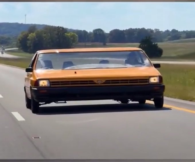 A gold-colored car driving down a highway, appearing to be the famously aerodynamic 'Whale Tail' or aero-coupe vehicle, possibly an Ford Probe or similar aerodynamic concept car.