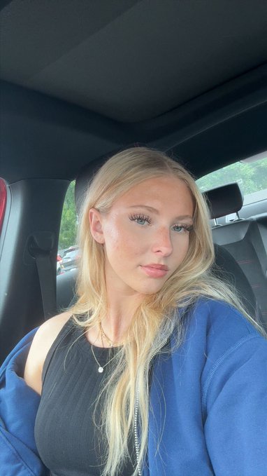 A blonde woman taking a selfie inside a car, wearing a black top and blue jacket/cardigan with a delicate gold necklace.