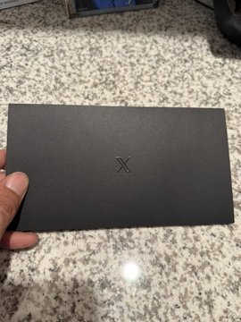 A person's hand holds a matte black box with an 'X' engraved on it, resting above a granite countertop.