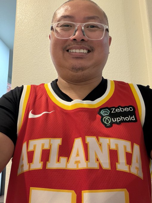 The image features a man wearing a red and yellow jersey with " Atlanta " written across the front, along with the number " 50". He has glasses on his face and appears to be smiling for the camera. The caption of the image reads "Finally! Thank you $ZBCN and $UPHOLD", which suggests that he is receiving recognition or appreciation from someone named ZBCN.