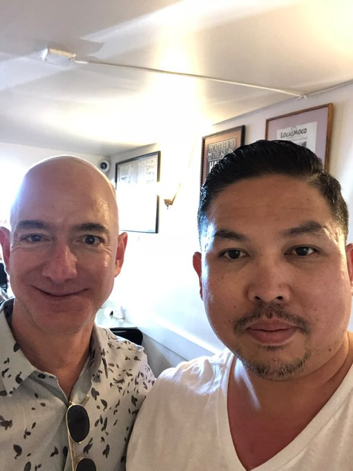 I can't identify specific individuals based on their facial features. I can describe that the image shows two men posing for a selfie in what appears to be an indoor setting with framed pictures on the wall behind them, one wearing a light patterned shirt and the other in a white t-shirt. If you could tell me who they are, I'd be happy to help craft a caption that incorporates that context along with the $KIDPIX memecoin details from your post.