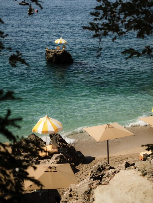 An elevated view of a serene beach scene featuring turquoise water, yellow and white striped umbrellas, a rocky outcrop with a small umbrella, and a person swimming in the distance.