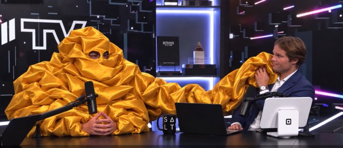 A man is wrapped in a yellow tarp while another man in a suit leans in to inspect him, in what appears to be a podcast studio setting. The scene relates to the caption by humorously portraying the man under the tarp as willingly complying with an absurd "security audit."