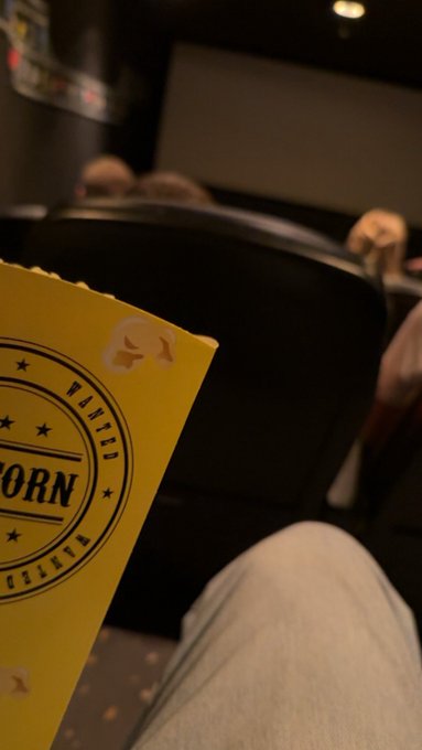 A person in a dark movie theater holds a yellow popcorn bag with a circular stamp design featuring stars and text including 'WANTED' and 'ORN', with their jeans visible in the foreground and other audience members ahead.