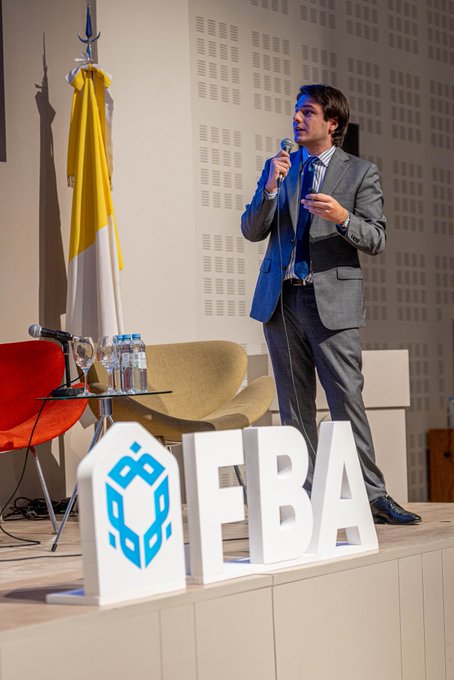 A man in a gray suit and blue tie speaks into a microphone on a stage, with a large "FBA" logo (featuring a blue cube) displayed prominently in front of him.