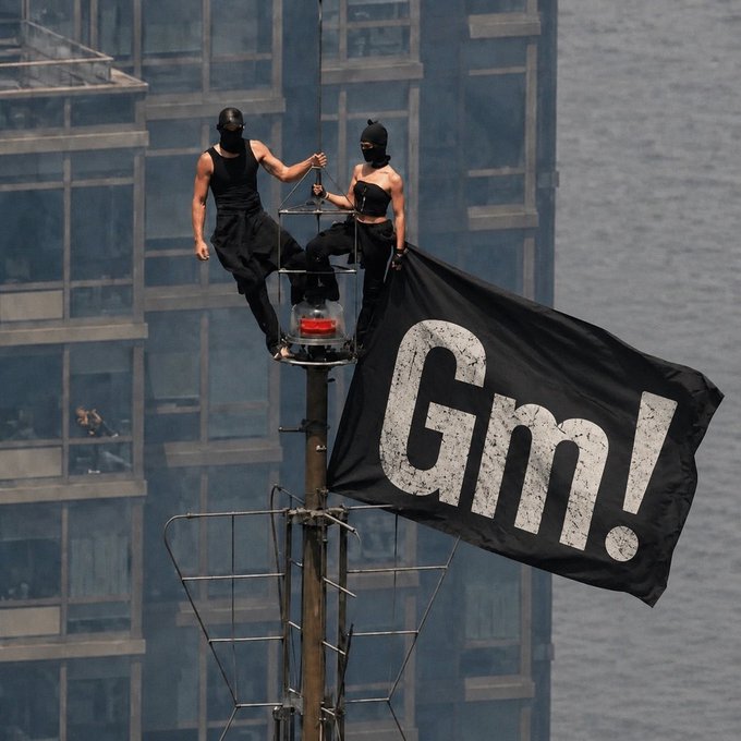 Two masked individuals in black attire perch atop a light pole displaying a large black flag with the white letters 'gm!', with a glass-fronted building in the background.
