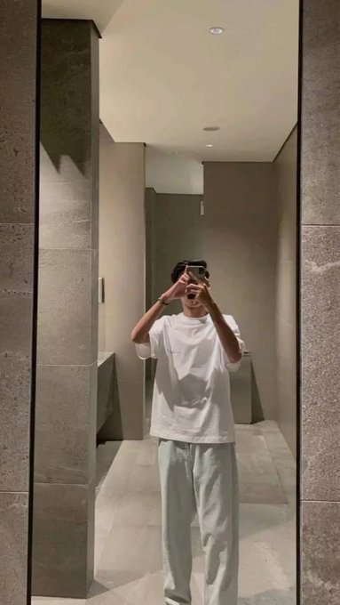 A person in a white t-shirt and light green pants takes a mirror selfie in a modern bathroom with gray tiled walls and a mirror reflecting their phone.