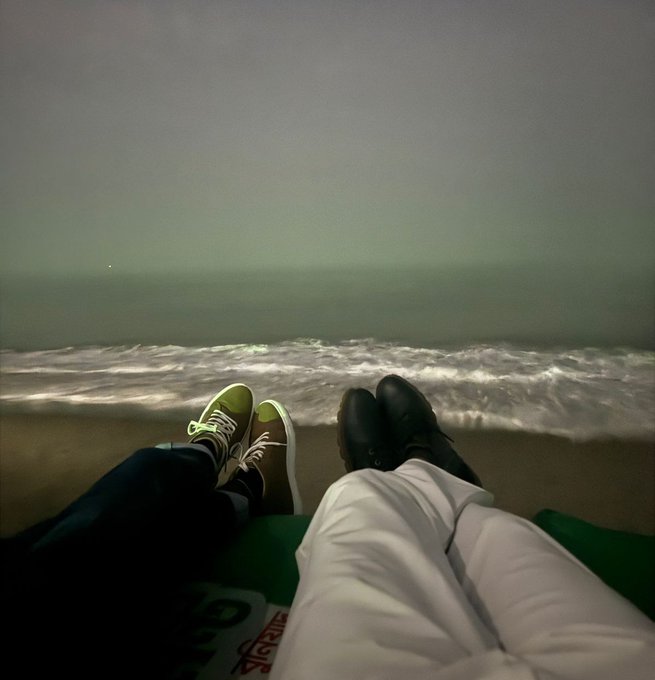 Two people sit on a beach at night with their feet up, one wearing green sneakers and the other in black shoes, with ocean waves visible in the background.