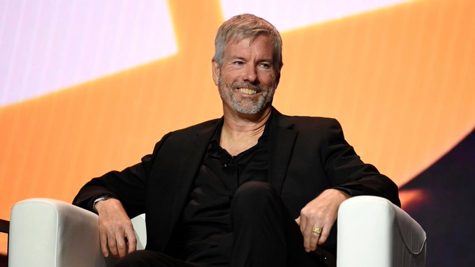 The image features a man sitting in an armchair, smiling at the camera while wearing a black suit. The background of the image shows a large screen with orange and white colors, which appears to be part of a stage or presentation setup. There is also text that reads "BREAKING: Michael Saylor just said ...", suggesting that this event might have been related to an important announcement or development in the cryptocurrency world.