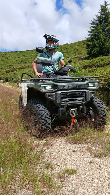A rider in a full-face helmet sits on a rugged Polaris ATV (with the winch and "Polaris" branding visible) parked on a mountain trail, surrounded by lush green hills and pine trees. Caption: "King of the mountain 👑 — Polaris power, no roads required."