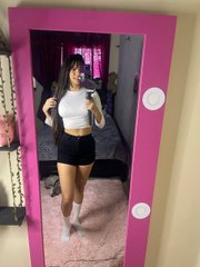 A mirror selfie of a woman wearing a white crop top and black shorts, standing in a room with a pink-framed mirror and a bed visible in the reflection.