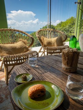 An outdoor dining setup with rattan chairs, a wooden table holding a plate with a single fried item and a glass, overlooking a scenic landscape under a partly cloudy sky.