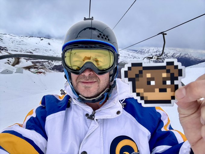 A snowboarder in a white helmet and yellow goggles takes a selfie on a snowy mountain while holding a pixelated dog sticker, embodying the "spiritual experience" of @doginaldogs by blending alpine adventure with meme culture.