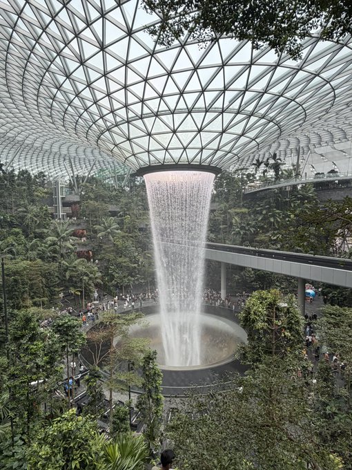 An interior view of the Jewel Changi Airport in Singapore, featuring the iconic Rain Vortex waterfall surrounded by lush indoor gardens under a geometric glass dome.