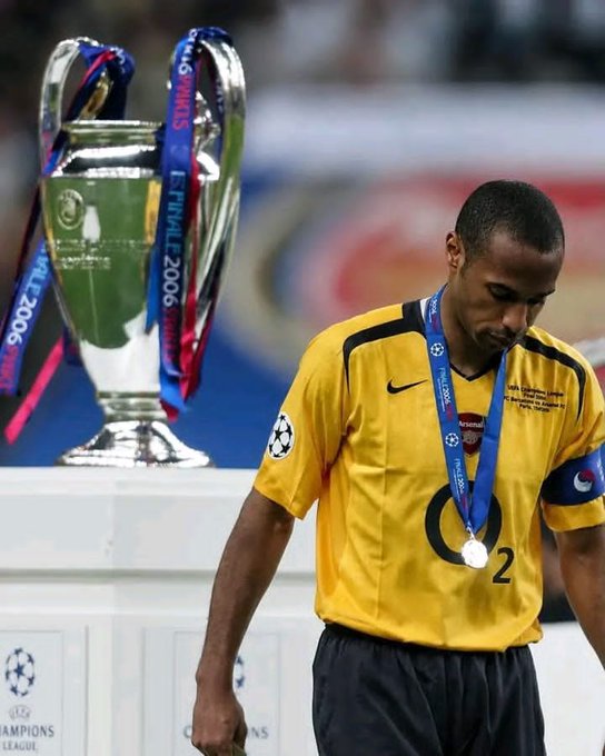 A soccer player in a yellow Arsenal jersey with a Champions League finalist's medal walks past the UEFA Champions League trophy in a moment of dejection.