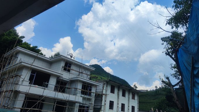 Photograph of a building under construction with scaffolding, set against a backdrop of green hills and a bright blue sky with fluffy clouds.