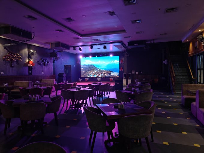 An interior view of a dimly lit lounge or event venue with purple and blue ambient lighting, featuring chairs and tables arranged around a stage with a large screen displaying a mountain landscape.