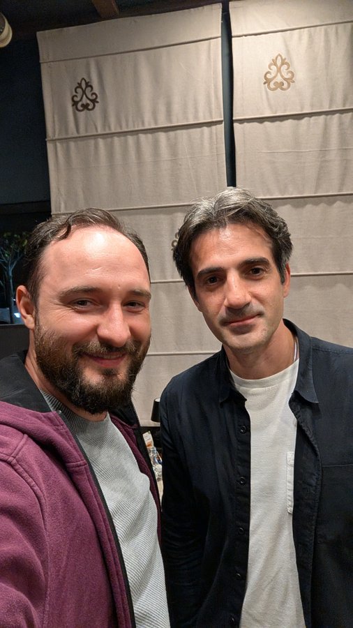Two men pose for a close-up selfie in front of decorative fabric panels with ornamental emblems. The casual, candid photo shares a personal moment alongside the "$VINE" post.