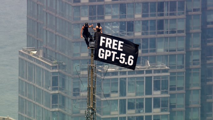 Two people perch atop an antenna in front of a glass skyscraper, unfurling a black banner that reads "FREE GPT-5.6" — a dramatic act of protest-cum-publicity referencing the awaited next-gen AI model.